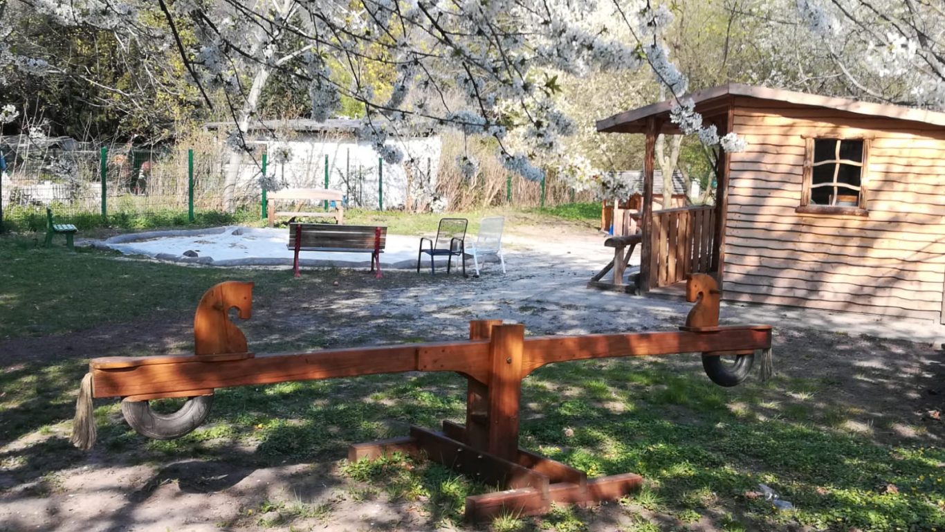 Ein Spielplatz mit einer Wippe und einem Holzhaus im Hintergrund, umgeben von Bäumen.