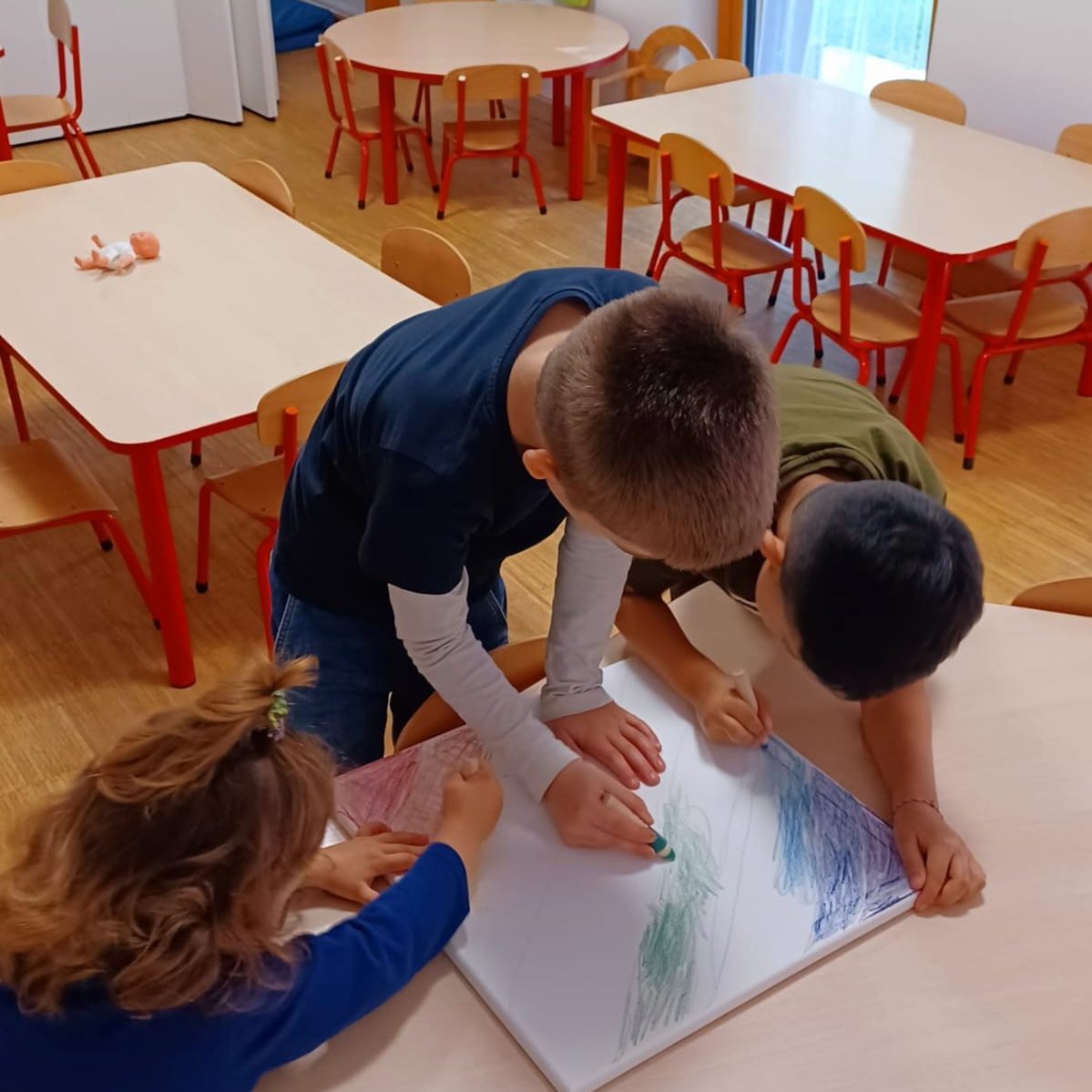 Drei Kinder zeichnen gemeinsam auf einem großen Blatt Papier in einem hellen Raum.