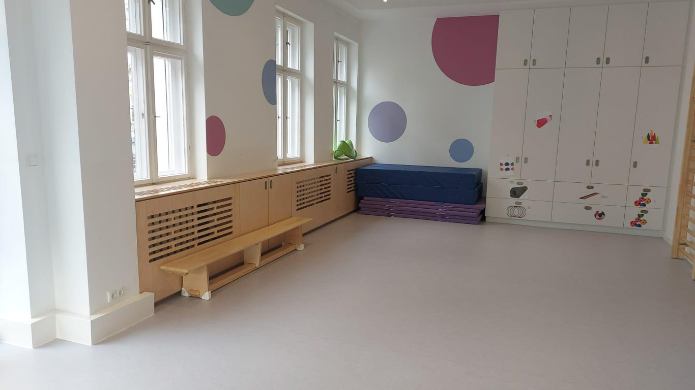 Heller Raum mit Holzschränken, einer Sitzbank und bunten Wandmalereien.