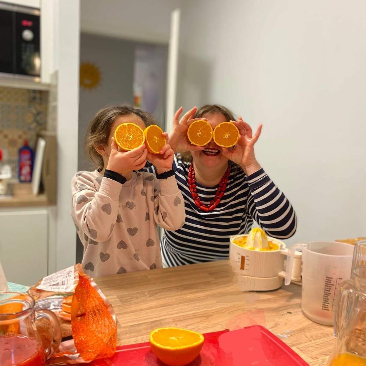 Zwei Personen halten Orangenscheiben vor ihre Augen und lächeln. Ein Tisch mit Küchenutensilien.