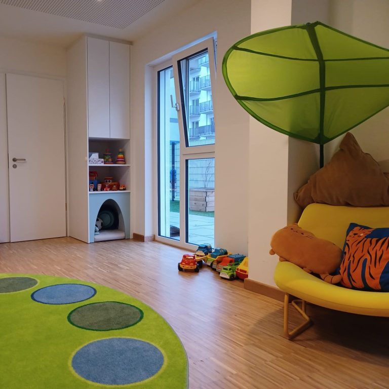 Spielzimmer mit buntem Teppich, grünem Sitzplatz und Spielzeug im Regal.