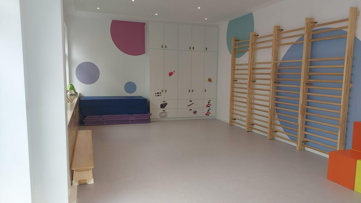 Großer Raum mit bunten Wandverzierungen, Turngeräten und einer Sitzbank.