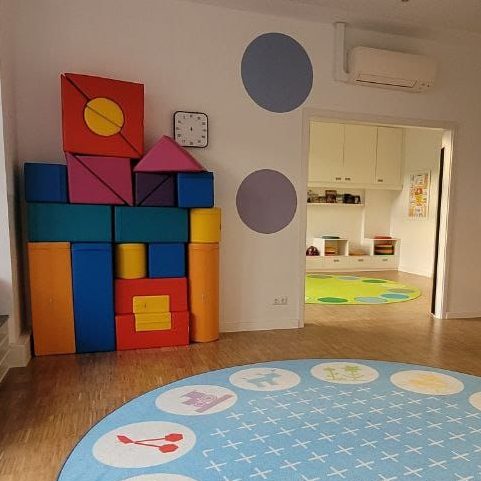 Bunte Bauklötze stehen an einer Wand in einem hellen Raum mit Teppich und Spielbereichen.